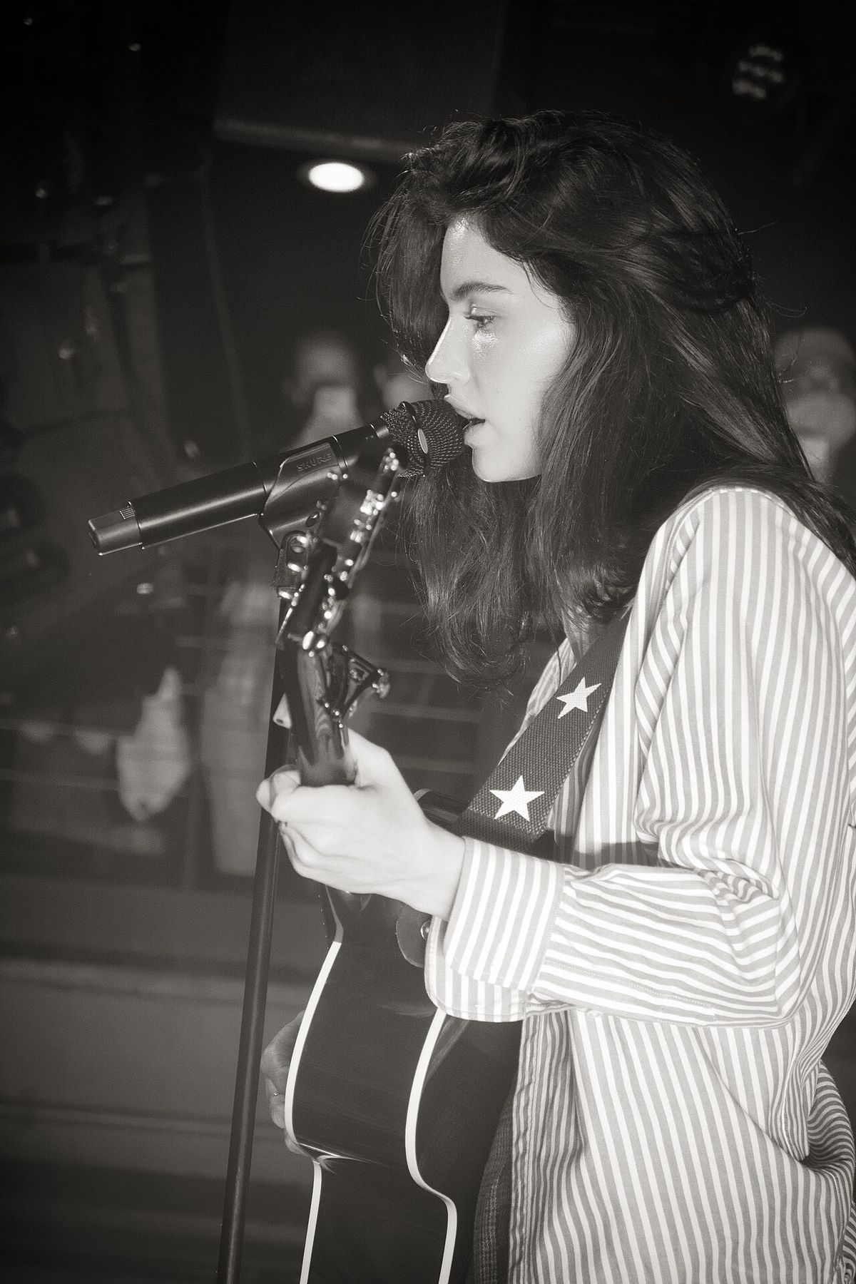 Gracie Abrams singing into a microphone with an acoustic guitar, in a black-and-white photo